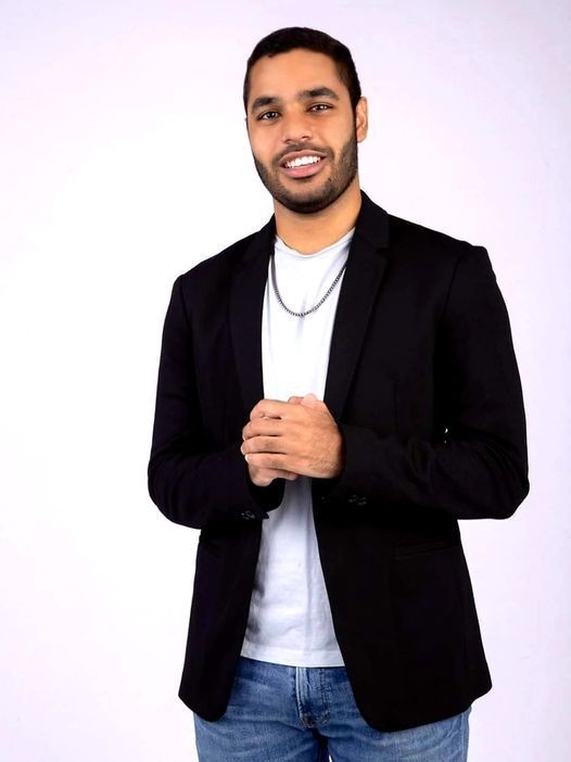 Smiling man in a black blazer and blue jeans standing against a plain white background.