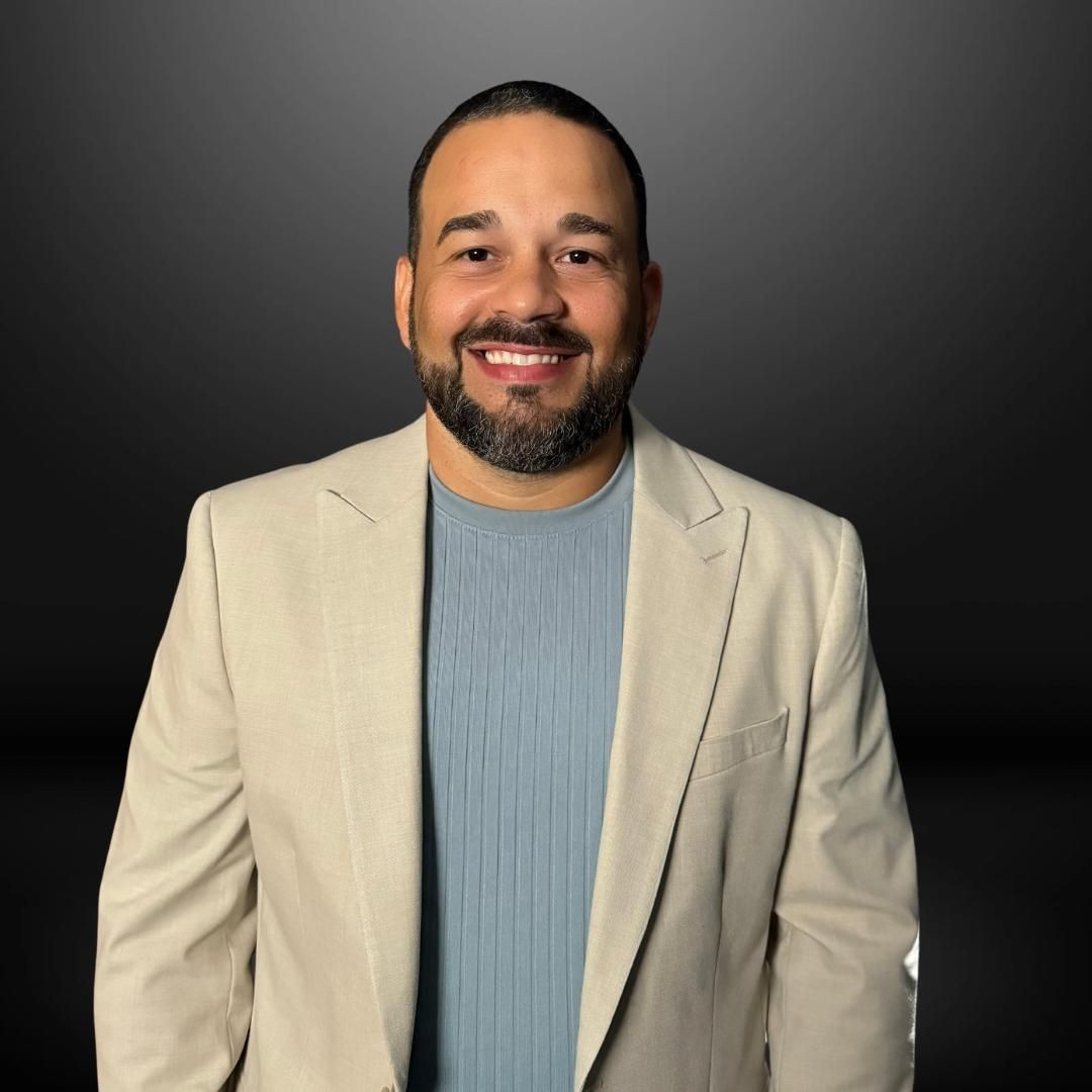 Man wearing a beige blazer over a blue ribbed shirt against a dark background.