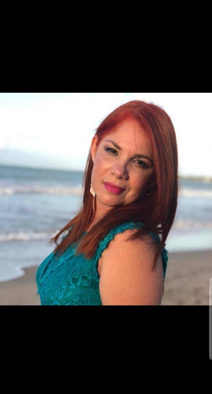 Person with red hair wearing a teal dress standing by the beach.