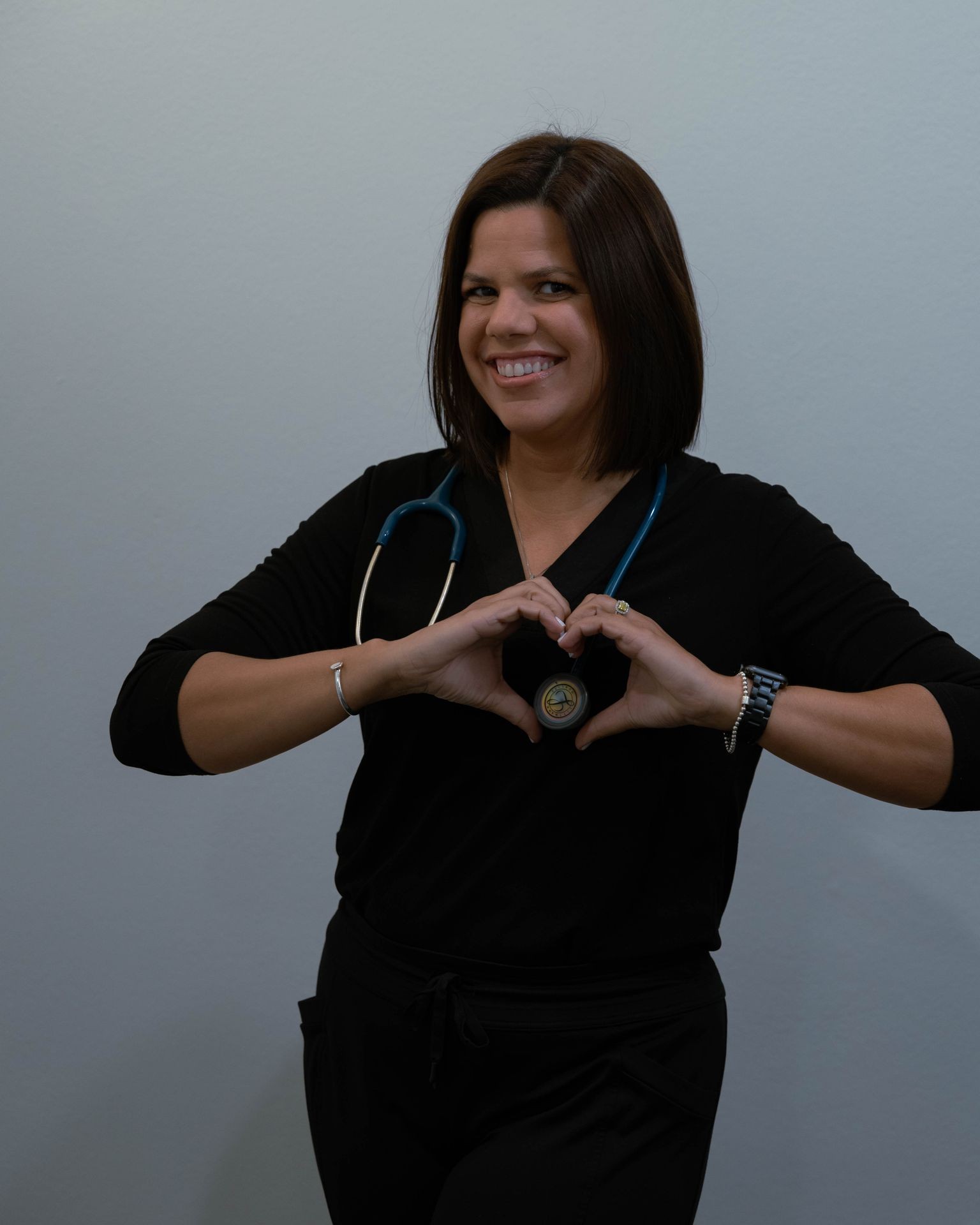 Person wearing a black outfit and stethoscope, making a heart shape with their hands.