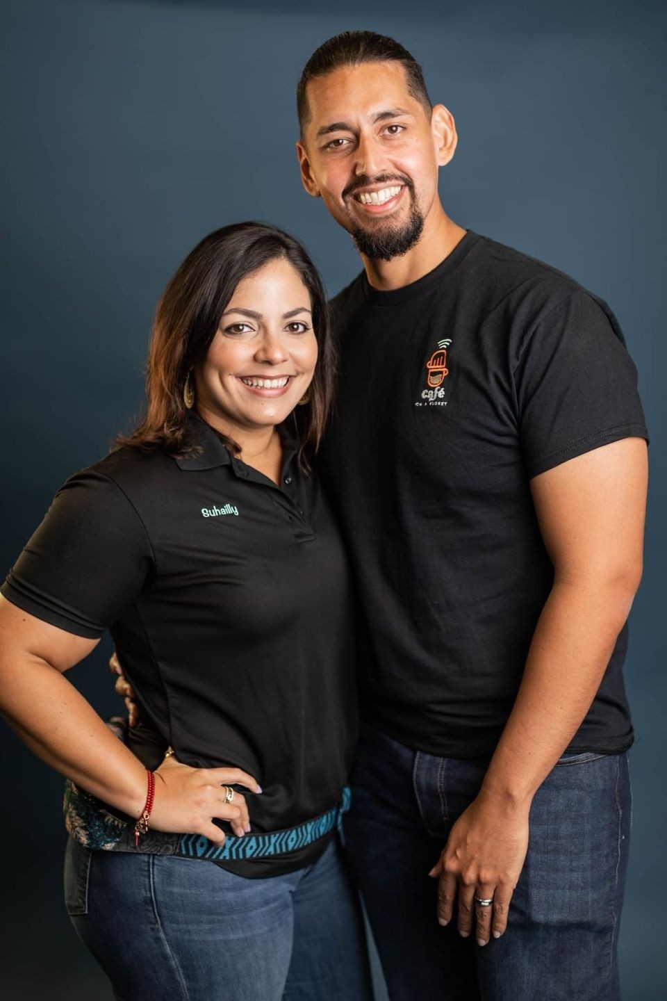 Two people standing close together, wearing black shirts and blue jeans, posing against a dark background.