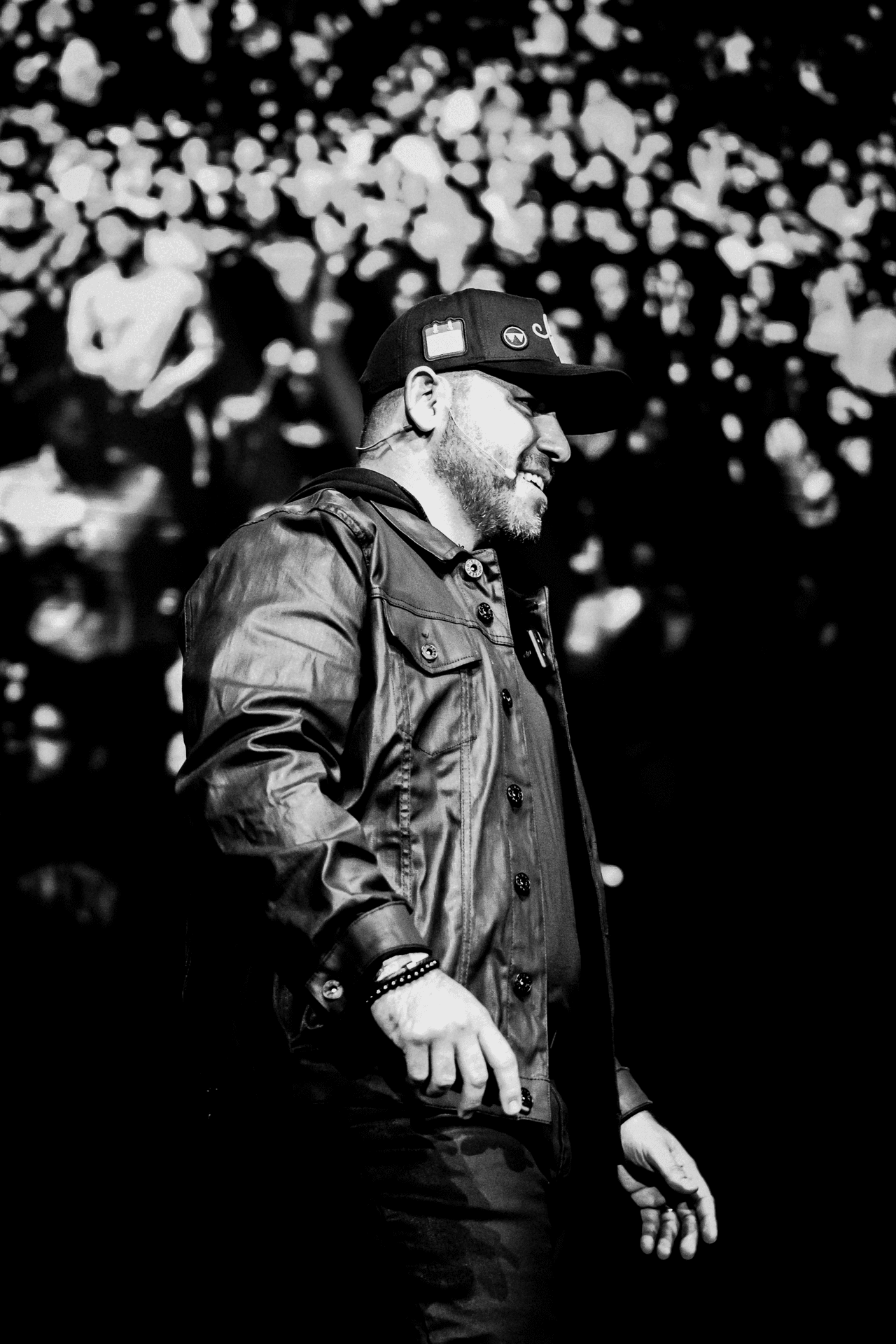 Person in a leather jacket and cap standing in front of a large crowd in black and white.