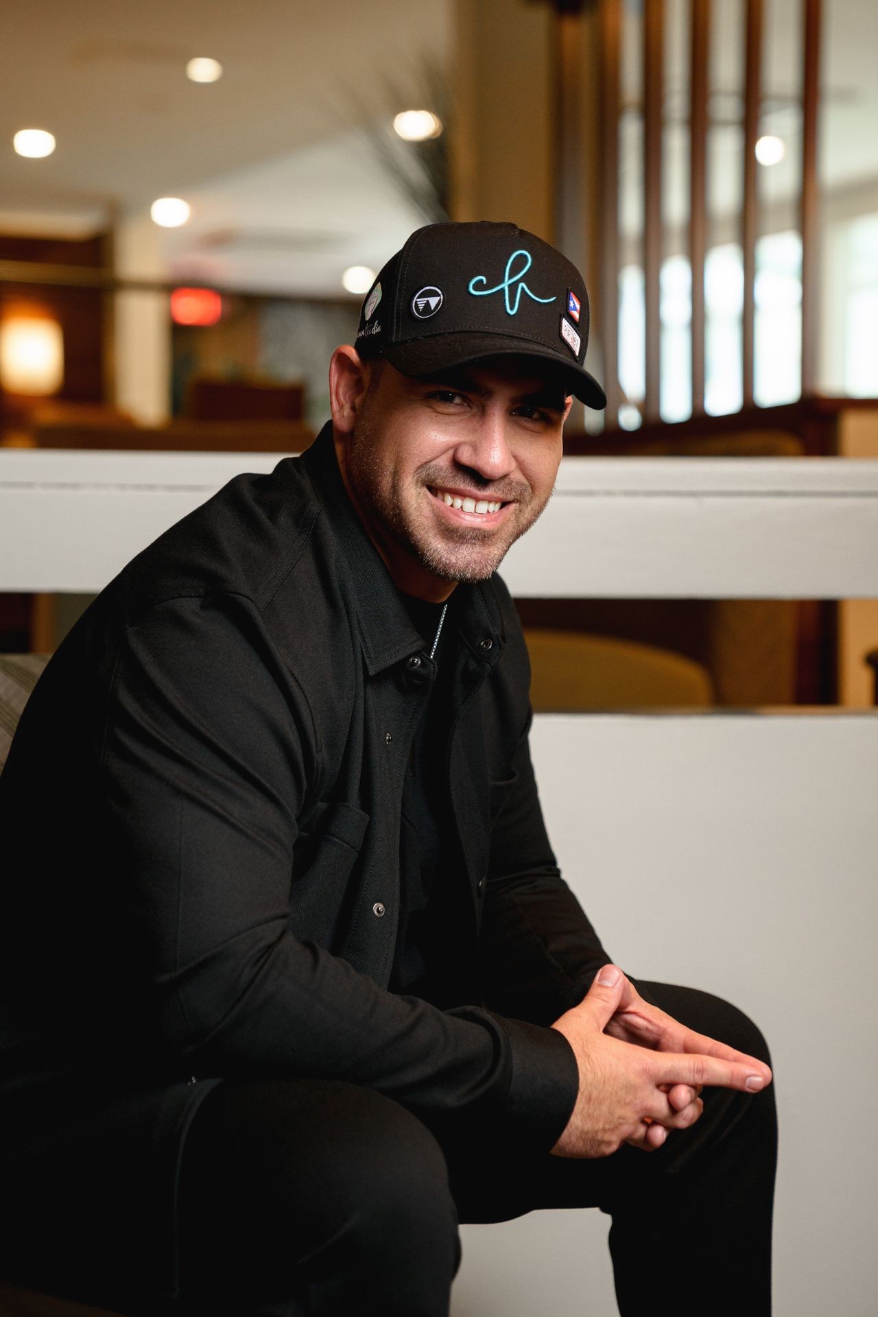 Man wearing a black hat and black shirt, sitting indoors and smiling at the camera.